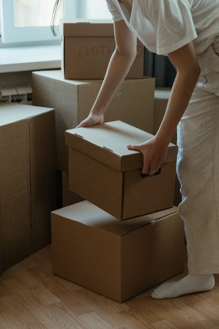 A man with curly black hair and a beard, dressed in a blue uniform, is seated on a wooden stool inside a room with a textured light grey wall. Around him are several packed cardboard boxes of various 